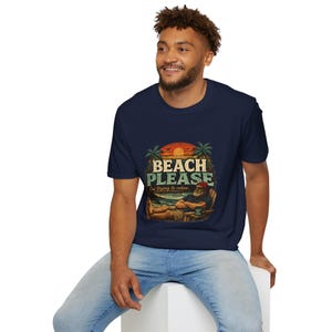 A smiling man wearing a navy blue t-shirt with a graphic design of a beach scene and the text "BEACH PLEASE" is sitting on a white stool.