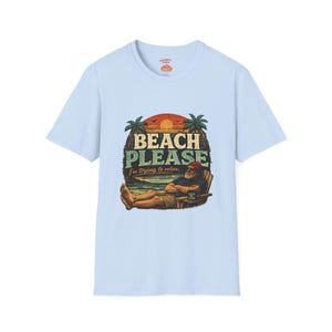 A light blue t-shirt with a graphic design depicting a man relaxing on a beach, with the text "BEACH PLEASE I'M DYING TO RELAX" printed on it.