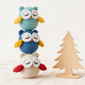 Może przedstawiać: Trzy szydełkowe sowy ułożone w stos, w odcieniach niebieskiego i kremowego, z żółtymi i czerwonymi akcentami. Każda sowa ma białe oczy i zamknięte oczy. Po prawej stronie stoi mała, brązowa, kartonowa choinka.