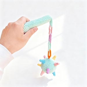 Könnte beinhalten: Ein gehäkeltes Babyrassel in Pastellfarben. Die Rassel hat einen hellblauen Griff und eine mehrfarbige Kette, die zu einem runden, stacheligen Ball führt. Der Ball hat verschiedenfarbige Stacheln. Die Rassel wird von einer Hand gehalten.