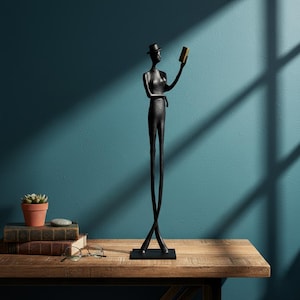 Peut inclure: Sculpture en métal noir représentant une silhouette élancée portant un chapeau et tenant un petit objet rectangulaire. La sculpture repose sur une base noire, avec de longues jambes croisées. Sur une table en bois, avec une plante en pot et des livres.