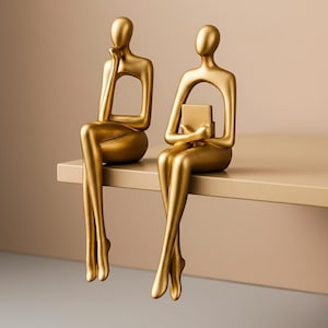 May include: Two gold-coloured abstract sculptures of seated figures. One figure rests their chin on their hand, the other holds a small rectangular object. The figures are seated on a shelf, with their legs dangling. The background is a neutral beige.