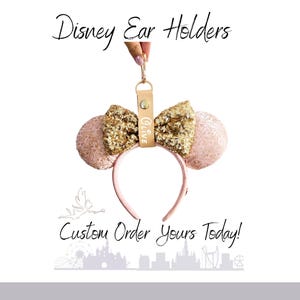 May include: A light pink headband with two round ears featuring a floral pattern and a central gold sequined bow. A beige leather strap with a gold snap closure and the name Olive printed on it hangs from the top. Text reads Disney Ear Holders.