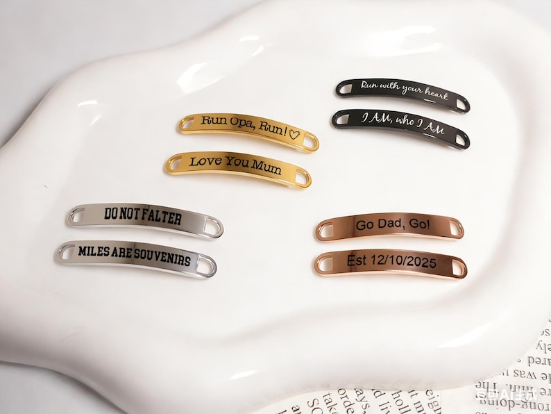 May include: A collection of curved metal tags in gold, silver, and rose gold. Each tag has engraved text, including phrases like "Run with your heart," "Love You Mum," and "Miles are souvenirs."