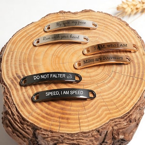 May include: A collection of running shoe tags in various metallic finishes, including silver, bronze, and black. Each tag features an inspirational phrase such as "My race, my pace" and "Speed, I am speed". The tags are displayed on a wooden surface.