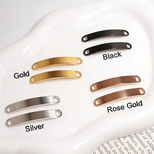 May include: An assortment of metal bar tags in gold, silver, black, and rose gold. The tags are curved with rounded ends and labeled with their respective colors. The tags are displayed on a white, oval-shaped dish.