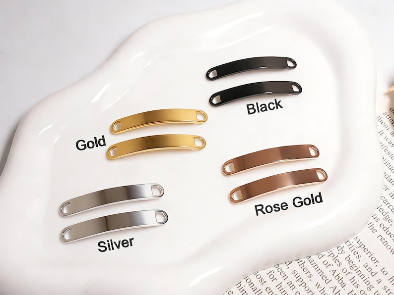 May include: An assortment of metal bar tags in gold, silver, black, and rose gold. The tags are curved with rounded ends and labeled with their respective colors. The tags are displayed on a white, oval-shaped dish.