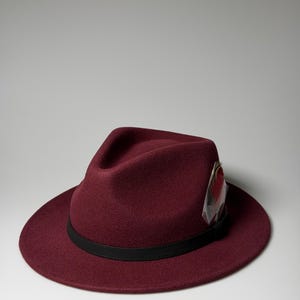 Może przedstawiać: Kapelusz typu fedora z ciemnobordowego filcu o strukturalnej koronie i szerokim rondzie. Kapelusz ozdobiony jest cienkim czarnym skórzanym paskiem otaczającym podstawę korony oraz pojedynczym ozdobnym piórem włożonym z boku paska.