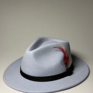 May include: A light blue fedora hat featuring a wide brim and a pinched crown. The hat is accented with a solid black band around the base and a decorative feather detail in shades of red, brown, and white tucked into the side of the band.