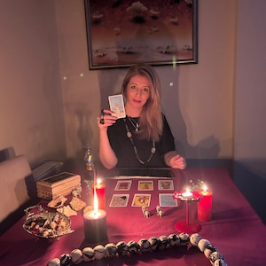 May include: A woman holding a tarot card, seated at a table with lit candles and tarot cards. A beaded necklace and a decorative bowl are on the table. A painting hangs on the wall behind her.