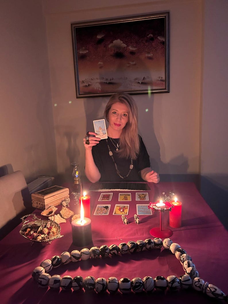 May include: A woman holding a tarot card, seated at a table with lit candles and tarot cards. A beaded necklace and a decorative bowl are on the table. A painting hangs on the wall behind her.