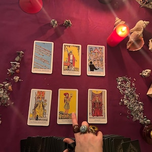 May include: A tarot card spread on a deep purple cloth, featuring illustrated cards with Turkish text. A hand adorned with rings gestures towards a card. Lit candles and decorative elements, including shells, are arranged around the cards.