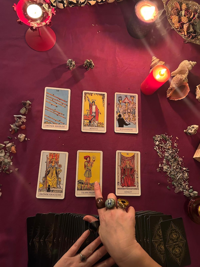 May include: A tarot card spread on a deep purple cloth, featuring illustrated cards with Turkish text. A hand adorned with rings gestures towards a card. Lit candles and decorative elements, including shells, are arranged around the cards.