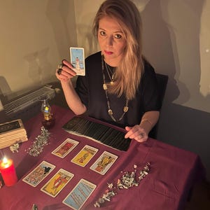 May include: A woman holding a tarot card, with a deck of cards and other tarot cards laid out on a claret table. Lit candles and decorative items are also on the table, creating a mystical atmosphere.