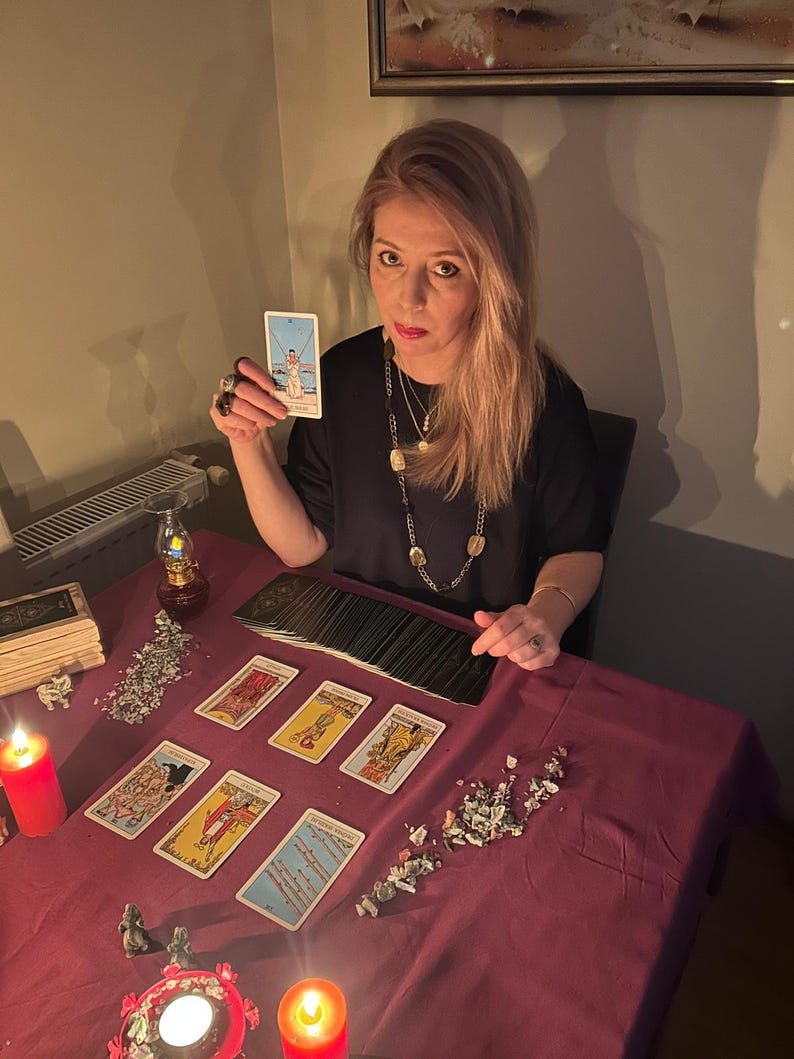 May include: A woman holding a tarot card, with a deck of cards and other tarot cards laid out on a claret table. Lit candles and decorative items are also on the table, creating a mystical atmosphere.