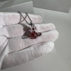 May include: Silver necklace with a red and silver pendant. The pendant has a red orb design with a silver cross and red accents. The necklace is displayed on a white surface, with a box in the background that says "Vivienne Westwood" and "Jewellery".