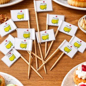 May include: Wooden toothpick flags with the Masters logo, featuring a yellow outline of the course and a red flag. The flags are scattered on a wooden table, near plates of pastries.