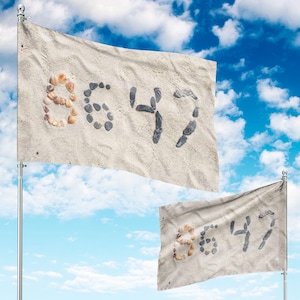 Könnte beinhalten: Zwei Flaggen mit sandfarbenem Hintergrund und den Zahlen "8647". Die Zahl "8" besteht aus Muscheln, während die anderen Zahlen aus dunkelgrauen Steinen bestehen. Die Flaggen sind vor einem blauen Himmel mit Wolken.