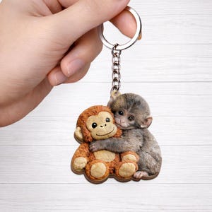 May include: A keychain featuring two hugging monkeys. One monkey is brown and the other is gray. The keychain has a silver chain and ring.