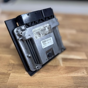 May include: A silver and black automotive electronic control unit (ECU) with a black plastic mounting bracket. The ECU has cooling fins and labels with text and barcodes. Likely for an Audi vehicle.