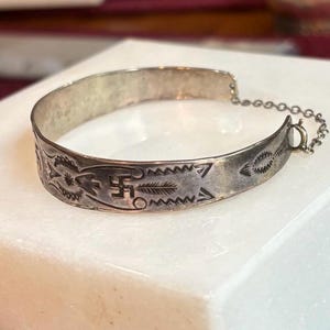 May include: A silver-toned metal cuff bracelet featuring engraved geometric patterns, including a central swastika symbol, arrow motifs, and serrated lines. The bracelet includes a thin attached metal safety chain with a small spring ring clasp.