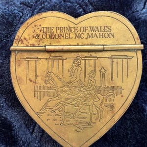 May include: A heart-shaped brass box featuring an engraved illustration of two figures on horseback. The text THE PRINCE OF WALES & COLONEL MC MAHON is etched across the top. The metal surface shows signs of age and oxidation with a visible hinge.