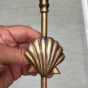 以下が含まれることがあります： 中央にホタテ貝のモチーフがあしらわれた真鍮製のキャンドルホルダー。円形の台座には葉の模様が浮き彫りにされており、上部にはキャンドルを立てるための円筒形のカップが付いています。金属は温かみのあるゴールドの仕上げです。