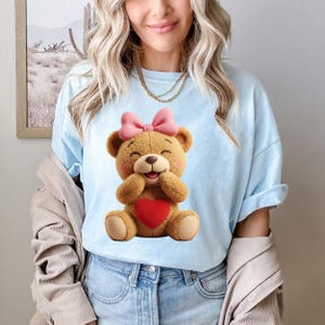 May include: Light blue t-shirt featuring a cartoon teddy bear with a pink bow, a red heart, and a smiling expression. The bear is hugging itself. The shirt is paired with blue jeans.