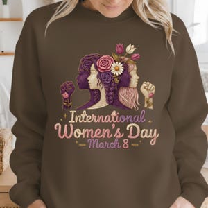 May include: Brown sweatshirt featuring a graphic for International Women's Day. The design includes three stylized female faces with floral accents and raised fists. The text "International Women's Day - March 8 -" is displayed in pink.