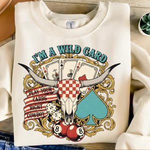 May include: Cream-colored sweatshirt with a graphic design featuring a bull skull, playing cards, dice, and the text "I'M A WILD CARD". The design includes the text "PLAY YOUR CARDS RIGHT COWBOY" on a red and white banner. The graphic also includes two red 8-balls.