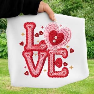 May include: White fabric with the word "LOVE" in red and pink knitted letters. A large pink knitted heart with a red heart in the center is above the "O". Small red hearts and gold stars are scattered around the word.
