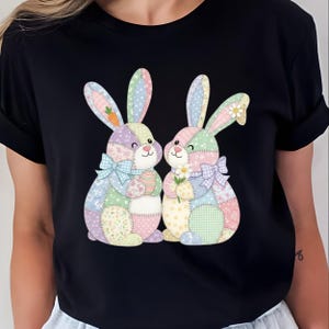 May include: Black t-shirt featuring a colorful graphic of two patchwork bunnies. One bunny holds an Easter egg, the other a daisy. The bunnies have pastel-colored ears and bows, perfect for Easter or spring.