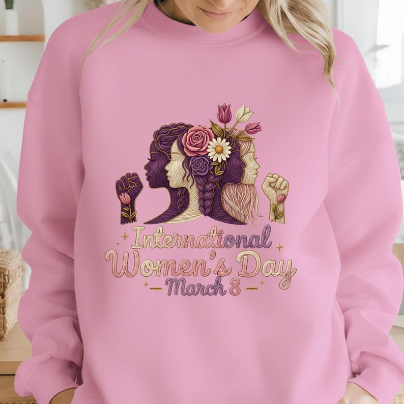 May include: Pink sweatshirt featuring a graphic for International Women's Day. The design includes three female profiles, flowers, and the text "International Women's Day - March 8". The artwork is in shades of purple, gold, and white.