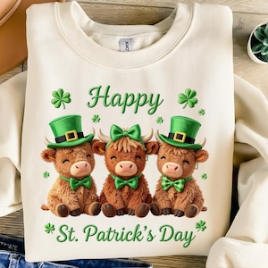 May include: Cream-coloured sweatshirt with three cartoon Highland cows. Each cow wears a green hat and bow tie, with a green bow on the middle cow. The words "Happy St. Patrick's Day" are written in green script with shamrock accents.