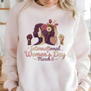 May include: White sweatshirt featuring a graphic celebrating International Women's Day. The design includes three female silhouettes, flowers, and the text "International Women's Day - March 8". The design is in shades of purple, pink, and white.