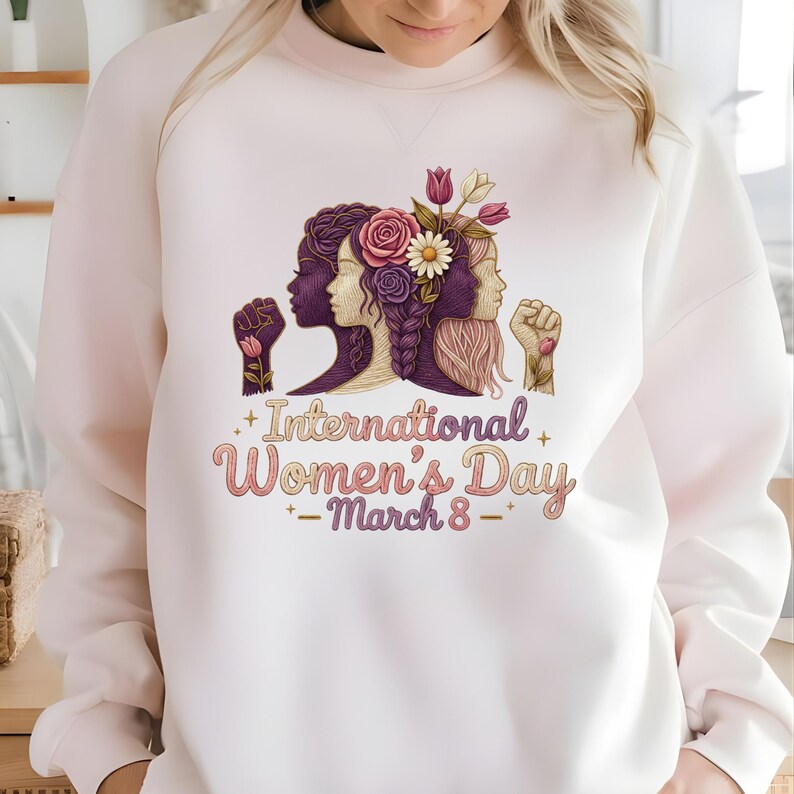 May include: White sweatshirt featuring a graphic celebrating International Women's Day. The design includes three female silhouettes, flowers, and the text "International Women's Day - March 8". The design is in shades of purple, pink, and white.