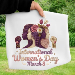 May include: White fabric featuring an illustration for International Women's Day. The design includes silhouettes of women with floral accents, fists, and the text "International Women's Day - March 8" in a decorative font. The image is held by a person.