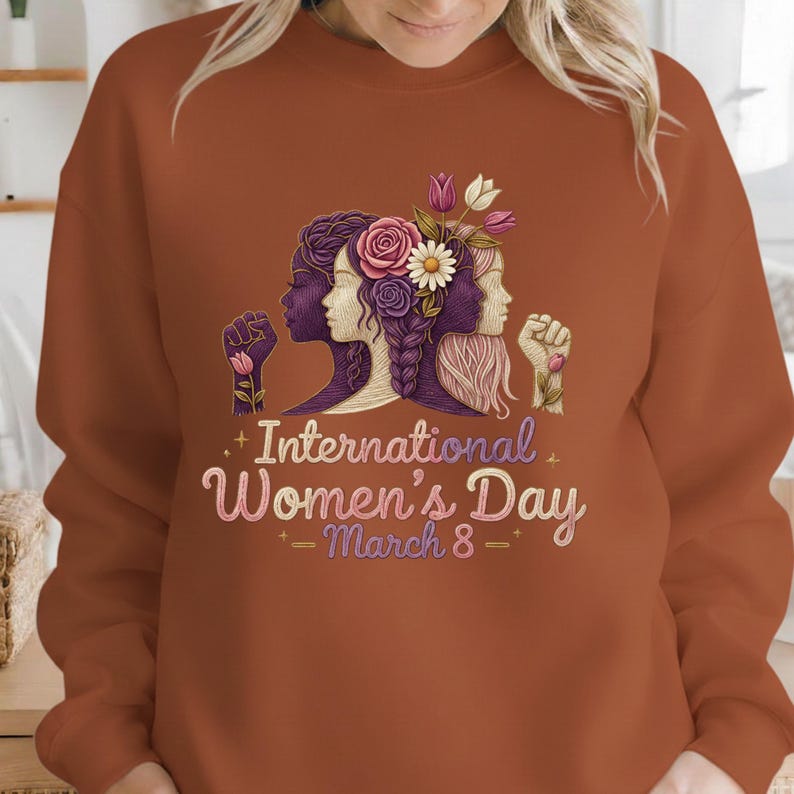 May include: A rust-colored sweatshirt featuring a graphic for International Women's Day. The design includes three female profiles, flowers, and the text "International Women's Day - March 8".