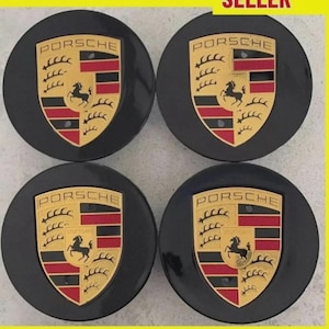 May include: Four black Porsche wheel center caps. Each cap features the Porsche emblem in gold, red, and black. The emblem includes the word "PORSCHE" and a horse.