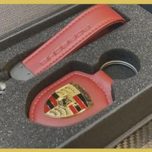 May include: A red leather keychain set with the Porsche logo, presented in a gray box. The set includes a key fob and a matching strap, both embossed with the brand name. The keychain has a silver metal ring.