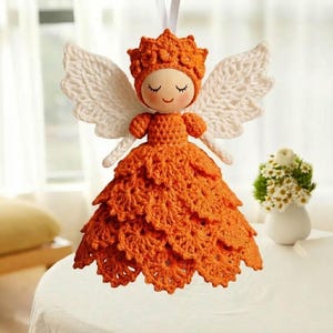以下が含まれることがあります： 白い翼と王冠が付いた、手作りのオレンジ色の編み物天使人形。 人形は、目を閉じて笑顔の表情をした木製の頭を持っています。 天使は白いリボンから吊り下げられており、休日の装飾に最適です。