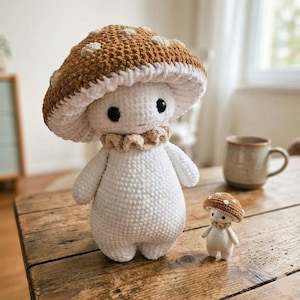 May include: Two crochet mushroom figures, one large and one small, sit on a wooden table. The larger mushroom has a white body, a brown cap with white spots, and a ruffled collar. The smaller mushroom has a similar design. A mug is in the background.