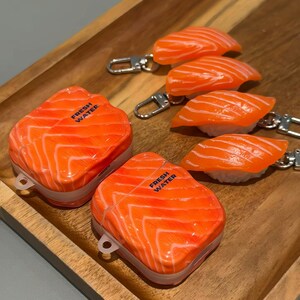 May include: A collection of salmon-shaped accessories, including keychains and cases. The keychains feature orange salmon nigiri, while the cases are shaped like salmon fillets. The text "FRESH WATER" is printed on the cases. All items are displayed on a wooden tray.