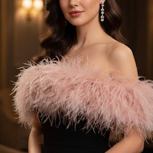 Puede incluir: Vestido negro sin tirantes con una boa de plumas rosa. El vestido tiene un corpiño ajustado y una falda fluida. La boa de plumas añade glamour al conjunto. Iluminación cálida y fondo borroso.