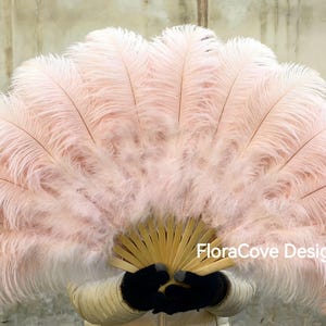 Può includere: Un grande ventaglio aperto fatto di morbide piume rosa chiaro. Il manico del ventaglio è in legno marrone chiaro. Il testo "FloraCove Design" è visibile sul ventaglio. Il ventaglio è tenuto da una persona con guanti neri.