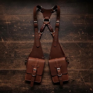 May include: A brown leather chest harness with two pouches and adjustable straps. The harness features metal buckles and rings, and is designed to be worn over the shoulders. The leather has a rich, dark brown color and a slightly distressed appearance.