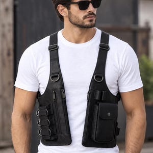 Può includere: Un gilet tattico nero a pettorina con scomparti multipli con cerniera, spallacci regolabili, anelli a D in metallo e una tasca frontale con patta. Il gilet è indossato sopra una t-shirt bianca e presenta vari passanti e componenti in metallo.