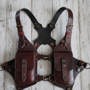 May include: A brown leather chest harness with two front pouches, adjustable straps, and metal hardware. The harness features a black leather back piece and is designed with decorative stitching.
