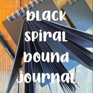 Könnte beinhalten: Schwarzes, spiralgebundenes Notizbuch mit einem Stift. Das Notizbuch ist geöffnet und zeigt seine schwarzen Seiten. Der Text "black spiral bound journal" ist auf dem Bild überlagert. Das Notizbuch liegt auf einer Holzoberfläche.