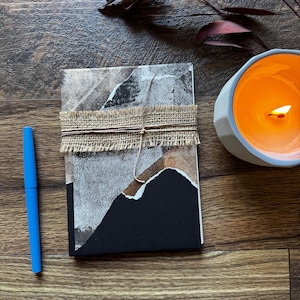 Pode incluir: Um diário feito à mão com um design abstrato em preto e branco, embrulhado com fita de serapilheira. Uma caneta azul e uma vela laranja acesa em um suporte cinza estão ao lado do diário. O fundo é uma superfície de madeira escura.